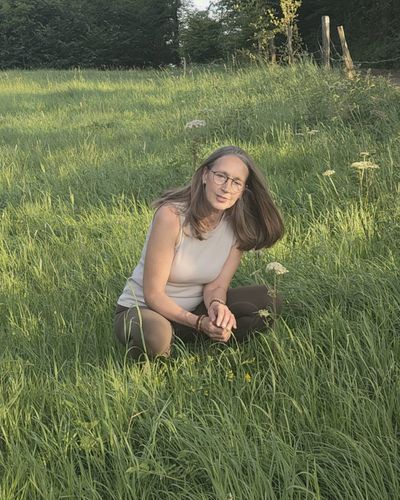 Heike kniet im hohen Gras und betrachtet blühende Wildkräuter am Wegesrand